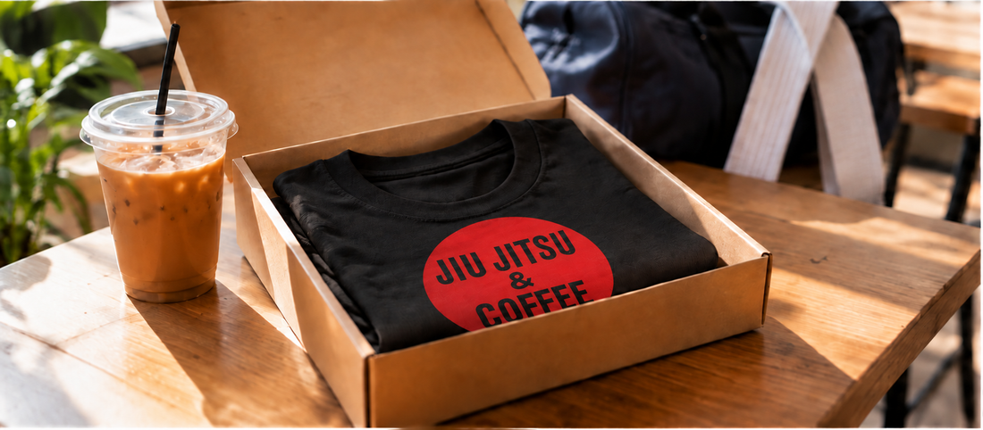 Gift box with a folded black Jiu Jitsu and Coffee t-shirt beside an iced coffee on a cafe table